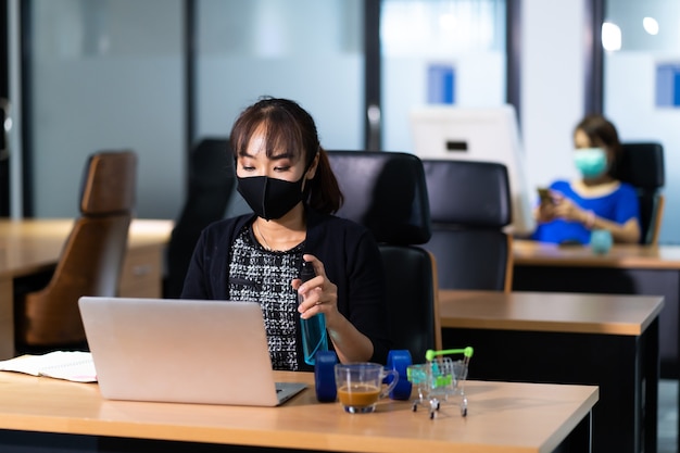 Jovem mulher asiática em spray desinfetante para policial em toalhetes limpando computador laptop na mesa de escritório contra o vírus corona covid-19