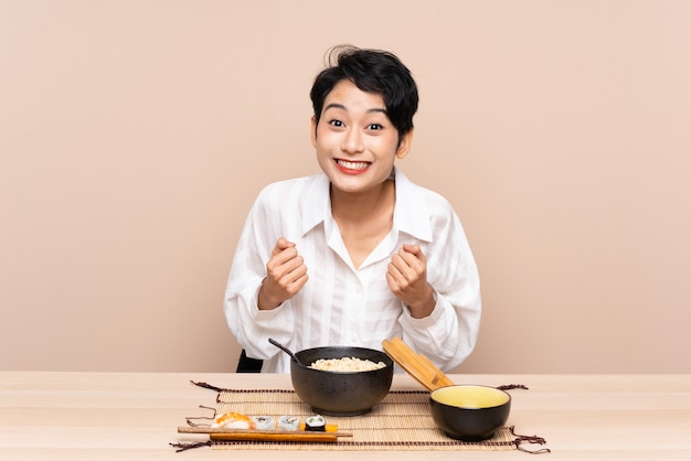 Jovem mulher asiática em uma mesa com uma tigela de macarrão e sushi comemorando uma vitória