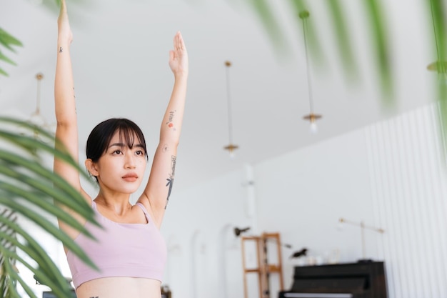 Jovem mulher asiática esportiva atraente praticando ioga fazendo exercício Virabhadrasana 1 meditando na pose de ioga Warrior One interior malhando em casa usando roupas esportivas Imagem com espaço de cópia