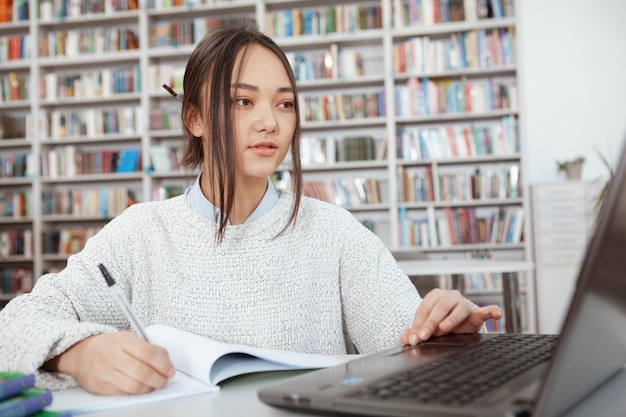 Jovem mulher asiática estudando na biblioteca da faculdade, usando seu laptop