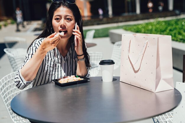 Jovem mulher asiática falando no telefone inteligente comendo salada na hora do almoço no parque da cidade, vivendo um estilo de vida saudável. Senhora feliz descansando conversando no celular após as compras do centro de stanford com sacos de café.