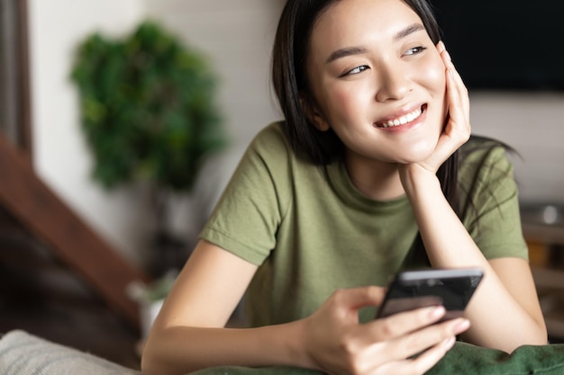 Jovem mulher asiática fazendo um pedido à espera de entrega no aplicativo para celular, sentada em casa e ...