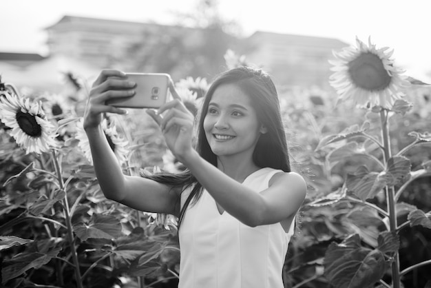 Jovem mulher asiática feliz sorrindo enquanto tira uma foto de selfie com o celular no campo de girassóis florescendo