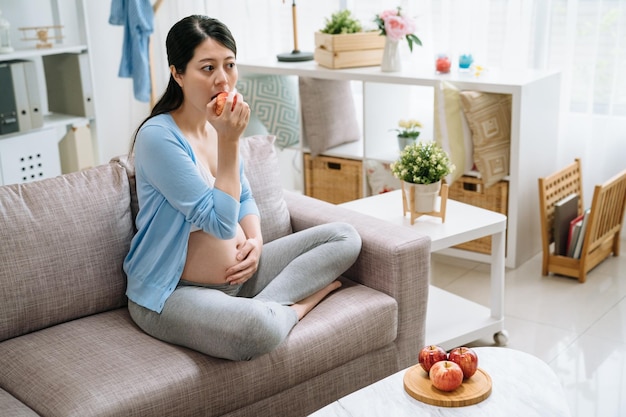 Jovem mulher asiática grávida segurando maçã vermelha com prato na mesa na sala de estar. mãe chinesa come comida vegana saudável sentada no sofá em casa. gravidez feminina com fome tendo energia comendo.