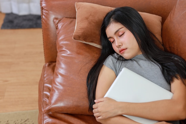 Jovem mulher asiática inteligente e ativa deitada e dormindo no sofá na sala de estar com tablet, estilo de vida e tecnologia.