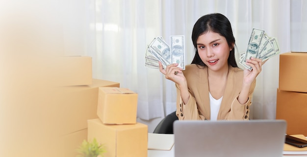 Jovem mulher asiática inteligente e feliz em um vestido casual, mostrando notas com emoção emocionante e trabalhando no computador para pedido de compra online e embalagem de caixa na mesa (novo conceito normal)