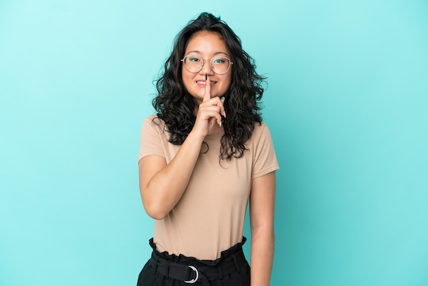 Jovem mulher asiática isolada em fundo azul mostrando sinal de silêncio gesto de colocar o dedo na boca