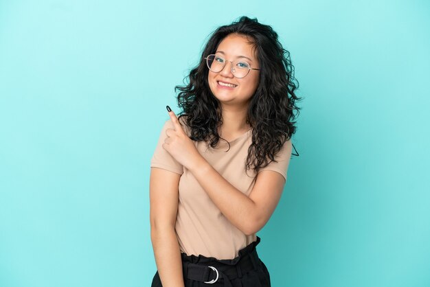 Jovem mulher asiática isolada em um fundo azul apontando para o lado para apresentar um produto