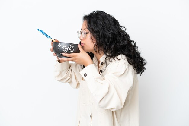 Jovem mulher asiática isolada no fundo branco segurando uma tigela de macarrão com pauzinhos e comendo