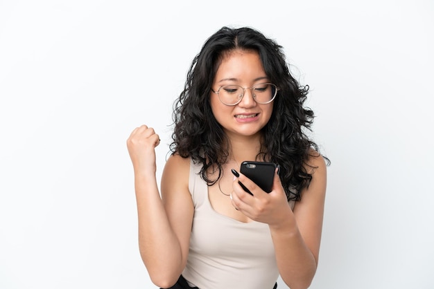 Jovem mulher asiática isolada no fundo branco usando telefone celular e fazendo gesto de vitória