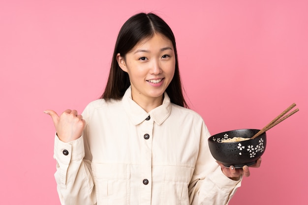 Jovem mulher asiática na parede rosa, apontando para o lado para apresentar um produto, mantendo uma tigela de macarrão com pauzinhos