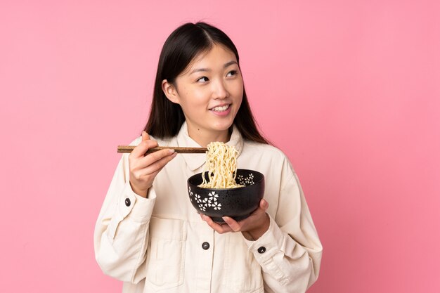 Jovem mulher asiática na parede rosa, segurando uma tigela de macarrão com pauzinhos e olhando para cima
