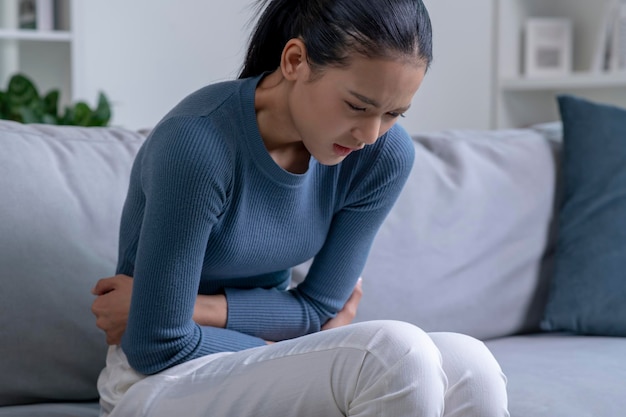 Jovem mulher asiática que sofre de forte dor abdominal enquanto está sentada no sofá em casa Mulher doente com dor de estômago
