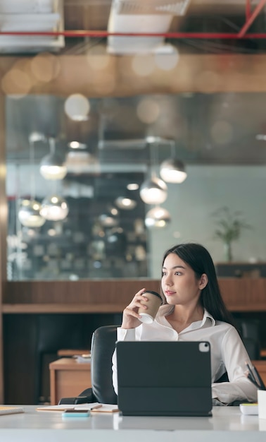 Jovem mulher asiática relaxando depois de trabalhar, segurando uma xícara de café e olhando para fora.