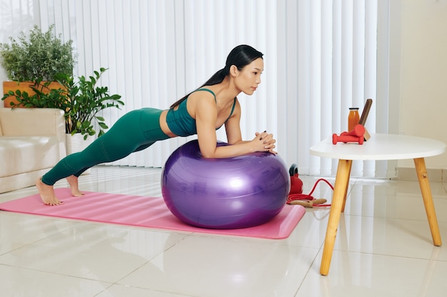 Jovem mulher asiática se equilibrando na bola de fitness ao seguir um treino de abdômen no computador tablet