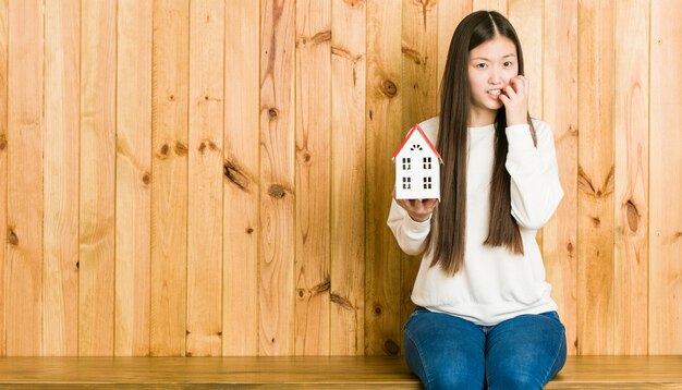 Jovem mulher asiática segurando as unhas roer um ícone de casa, nervoso e muito ansioso.