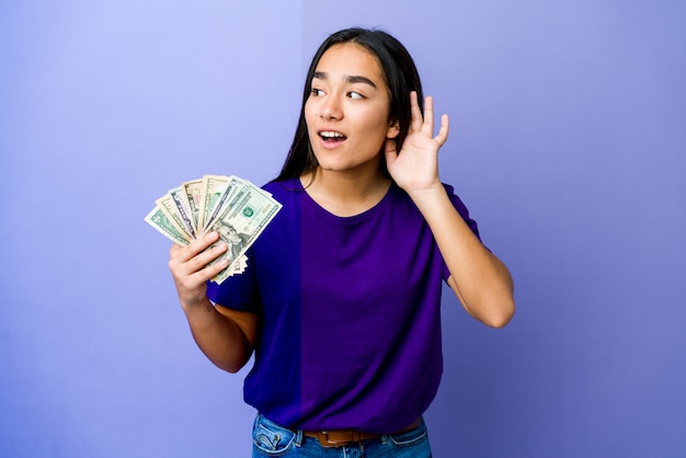 Jovem mulher asiática segurando dinheiro isolado na parede roxa, tentando ouvir uma fofoca.