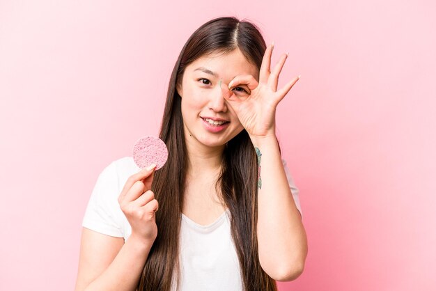 Jovem mulher asiática segurando esponja facial isolada no fundo rosa animado mantendo o gesto okey no olho