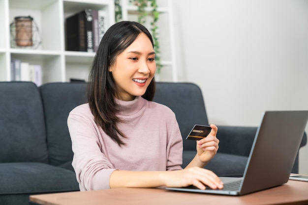 Jovem mulher asiática, segurando o cartão de crédito e usando o laptop para fazer compras on-line com pagamento no banco na internet.