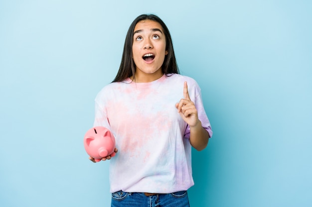 Jovem mulher asiática segurando um banco rosa sobre uma parede isolada apontando para cima com a boca aberta.