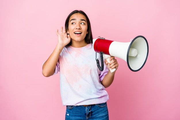 Jovem mulher asiática segurando um megafone isolado na rosa, tentando ouvir uma fofoca.