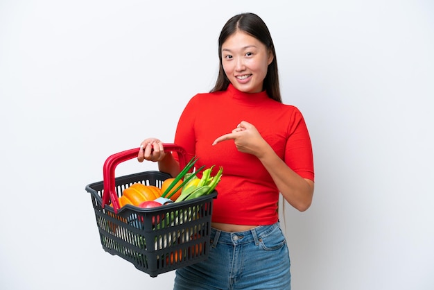 Jovem mulher asiática segurando uma cesta de compras cheia de comida isolada no fundo branco apontando para o lado para apresentar um produto