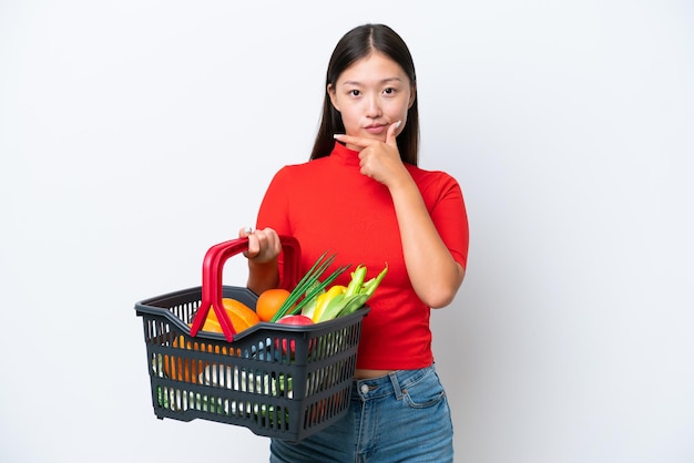 Jovem mulher asiática segurando uma cesta de compras cheia de comida isolada no pensamento de fundo branco