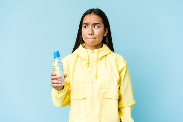 Jovem mulher asiática segurando uma garrafa de água isolada na parede azul confusa, sente-se em dúvida e insegura.