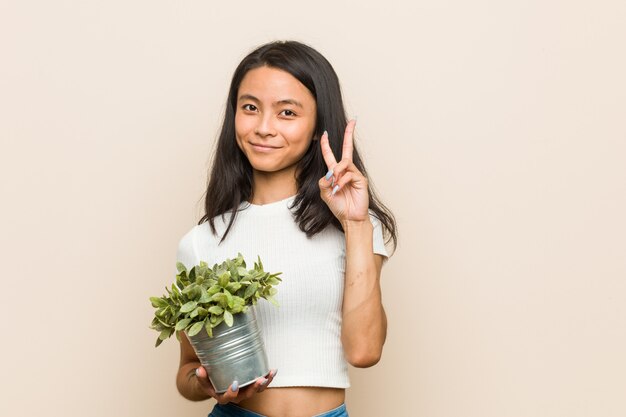 Jovem mulher asiática segurando uma planta mostrando o número dois com os dedos.