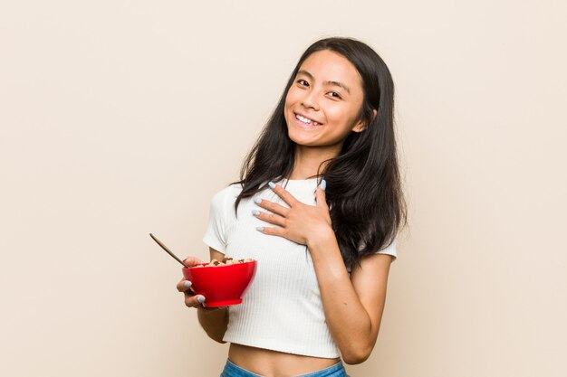 Jovem mulher asiática segurando uma tigela de cereais ri alto, mantendo a mão no peito.