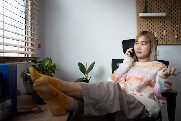 Jovem mulher asiática sentada em uma cadeira confortável na sala de estar e falando no celular.