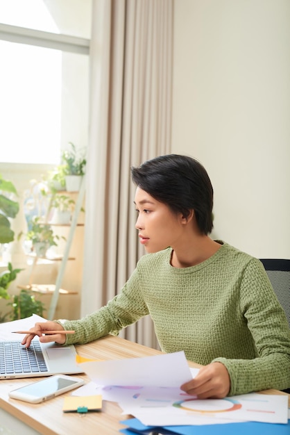 Jovem mulher asiática sentada na mesa do escritório olhando para a tela do computador portátil