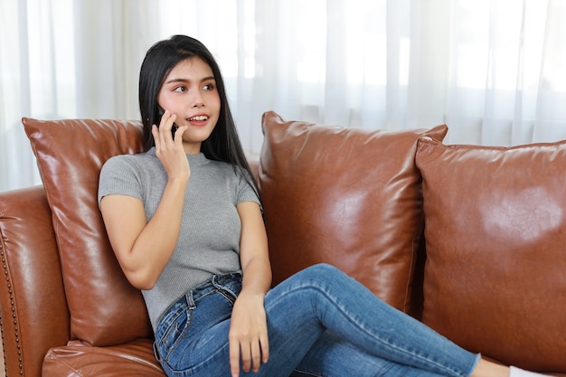 Jovem mulher asiática sentada no sofá da sala, usando o smartphone com uma cara feliz