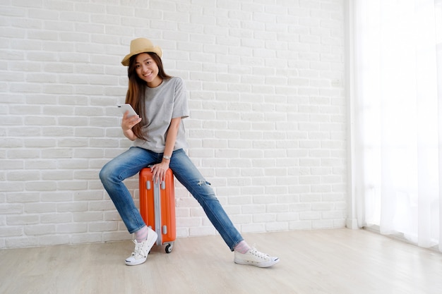 Jovem mulher asiática sentado na bagagem, segurando o smartphone na sala branca, turista adolescente feliz em casual, sente-se na bagagem de viagem, mala, bagagem pronta para viagem de férias de férias