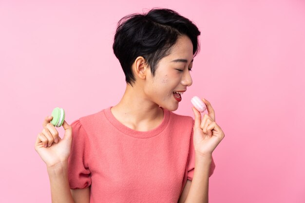 Jovem mulher asiática sobre parede rosa isolada segurando macarons franceses coloridos e comê-lo