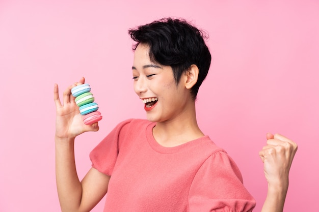 Jovem mulher asiática sobre parede rosa isolada segurando macarons franceses coloridos e comemorando uma vitória