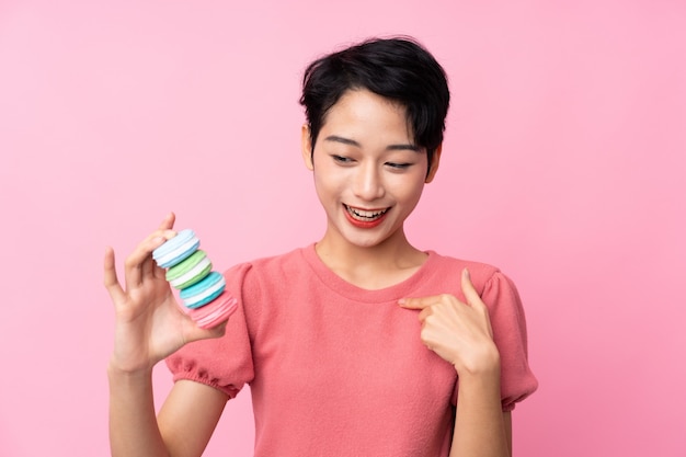 Jovem mulher asiática sobre parede rosa isolada segurando macarons franceses coloridos e comemorando uma vitória