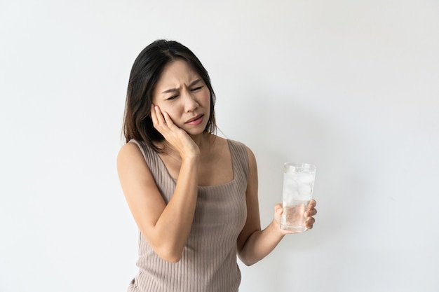 Jovem mulher asiática sofrendo de dor de dente e mão segurando um copo de água fria com gelo. Menina bebendo bebida gelada, copo cheio de cubos de gelo e sente dor de dente Conceito de saúde.