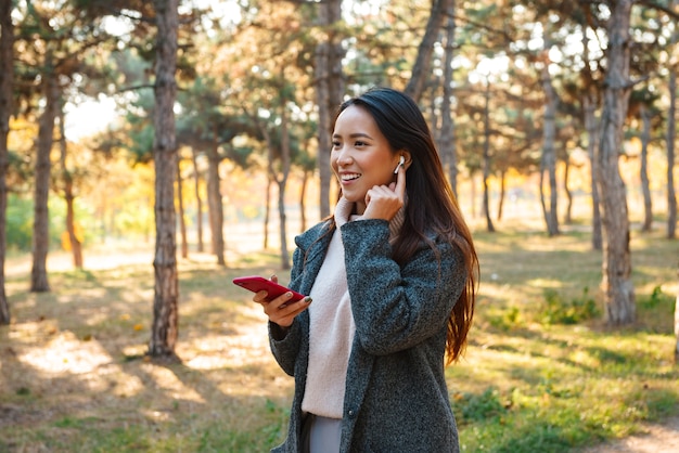 Jovem mulher asiática sorridente, vestindo um casaco, caminhando ao ar livre no parque, ouvindo música com fones de ouvido, segurando um telefone celular