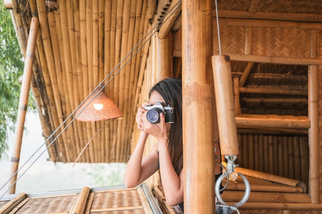 Jovem mulher asiática tirando foto com câmera retrô pela janela no resort de cabana de madeira