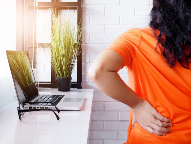 Jovem mulher asiática trabalhando com computador portátil e sentado na cadeira e sofrendo dores nas costas e coluna vertebral doloridas e dor de cintura, conceito de saúde e dores no corpo.