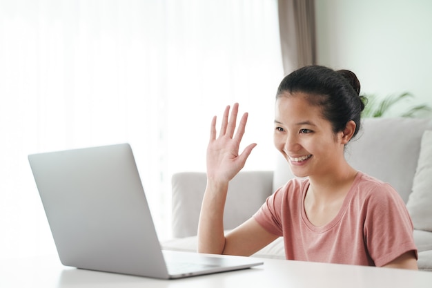 Jovem mulher asiática usando computador portátil para videoconferência online, acenando com a mão, fazendo um gesto de olá na sala de estar.