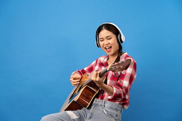 Foto jovem mulher asiática usando fone de ouvido e camisa vermelha de guingão com penteado de trança enquanto toca guitarra