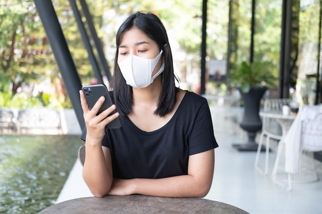 Jovem mulher asiática usando máscara médica usando smartphone em uma cafeteria Trabalho à distância social