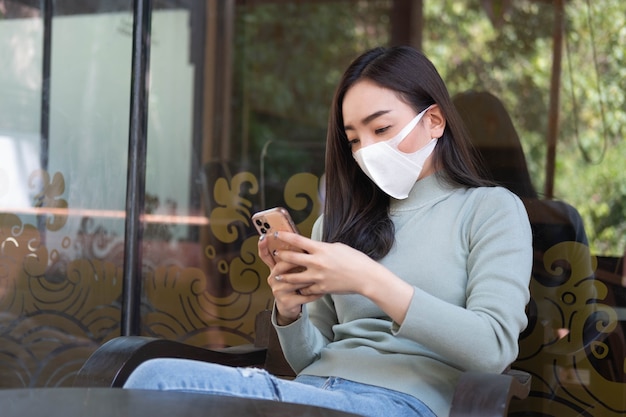 Jovem mulher asiática usando máscara médica usando smartphone em uma cafeteria trabalho à distância social