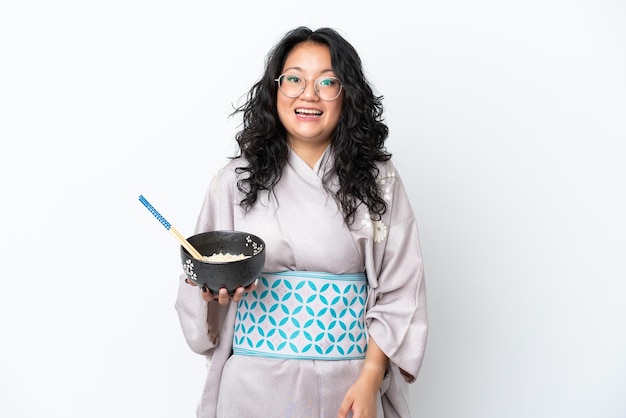 Jovem mulher asiática usando quimono isolado no fundo branco com expressão facial de surpresa e choque, segurando uma tigela de macarrão com pauzinhos