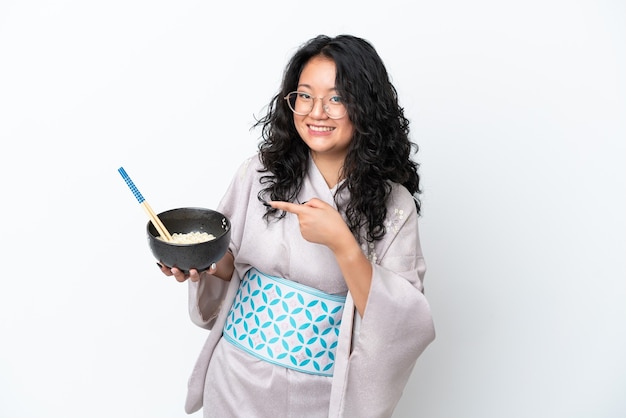 Jovem mulher asiática usando quimono isolado no fundo branco e apontando-o enquanto segura uma tigela de macarrão com pauzinhos
