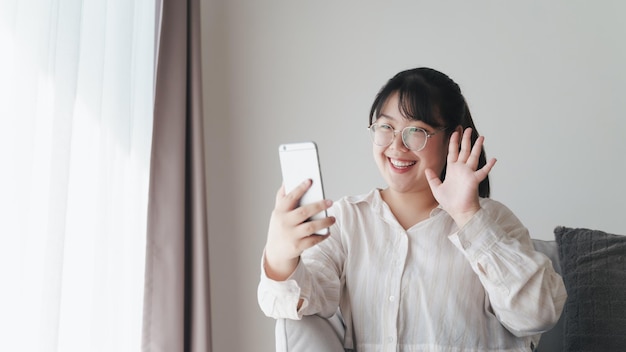 Jovem mulher asiática usando smartphone para videoconferência online acenando com a mão fazendo gesto de olá