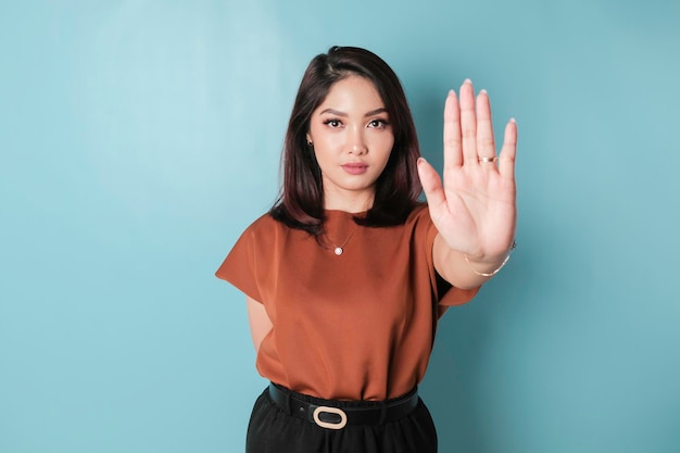 Jovem mulher asiática vestindo camisa marrom sobre fundo azul isolado fazendo sinal de pare com a palma da mão