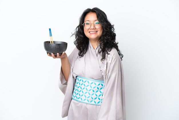 Jovem mulher asiática vestindo quimono isolado no fundo branco com uma expressão feliz, segurando uma tigela de macarrão com pauzinhos